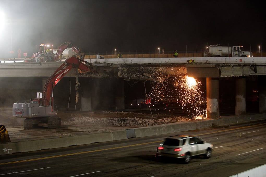 While a vehicle speeds south, the hoe ram punches at the north façade. Water trucks on the bridge spray water on the work area to reduce dust. Concrete from the bridge will be crushed on the project crusher sites and reused in the project. Photo by Peter Watkinson.