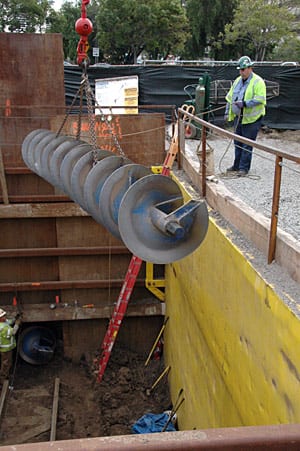 A worker guides a 24-foot-long auger as a crane lifts it from a work area north of Wilshire Bl. Another auger at the lower left waits to be removed. Chain rigging is used due to the sharp edges and irregular shape of the auger.